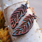 Colorful feather-shaped earrings on a white surface with natural elements.