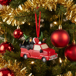 Christmas ornament of a black bear in a red truck with a tree, on a white background.