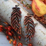 Feather-shaped earrings on a wooden log with autumn leaves and berries.