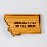 Wooden state-shaped sign with 'Montana Sucks (Tell Your Friends)' text on a white background