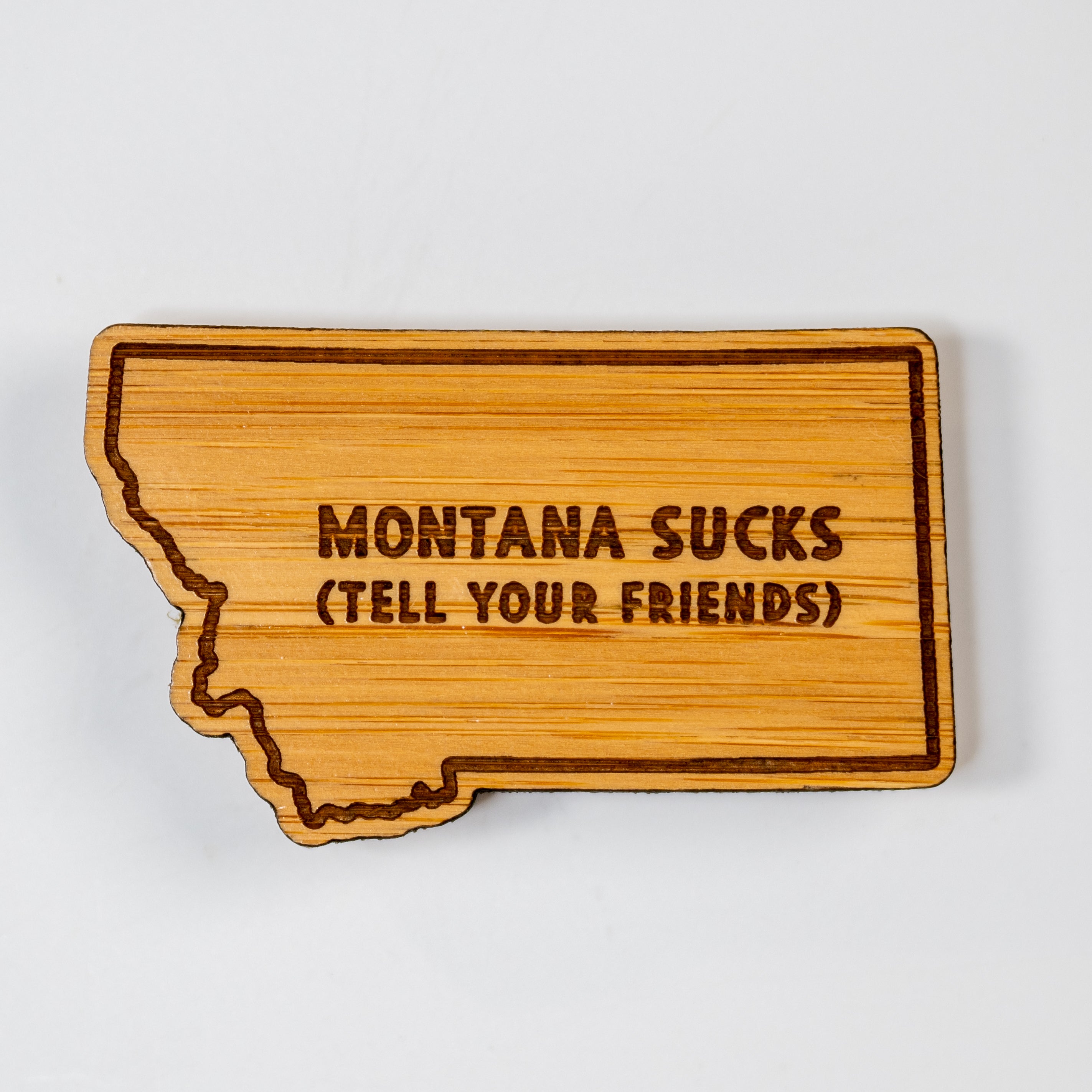 Wooden state-shaped sign with 'Montana Sucks (Tell Your Friends)' text on a white background