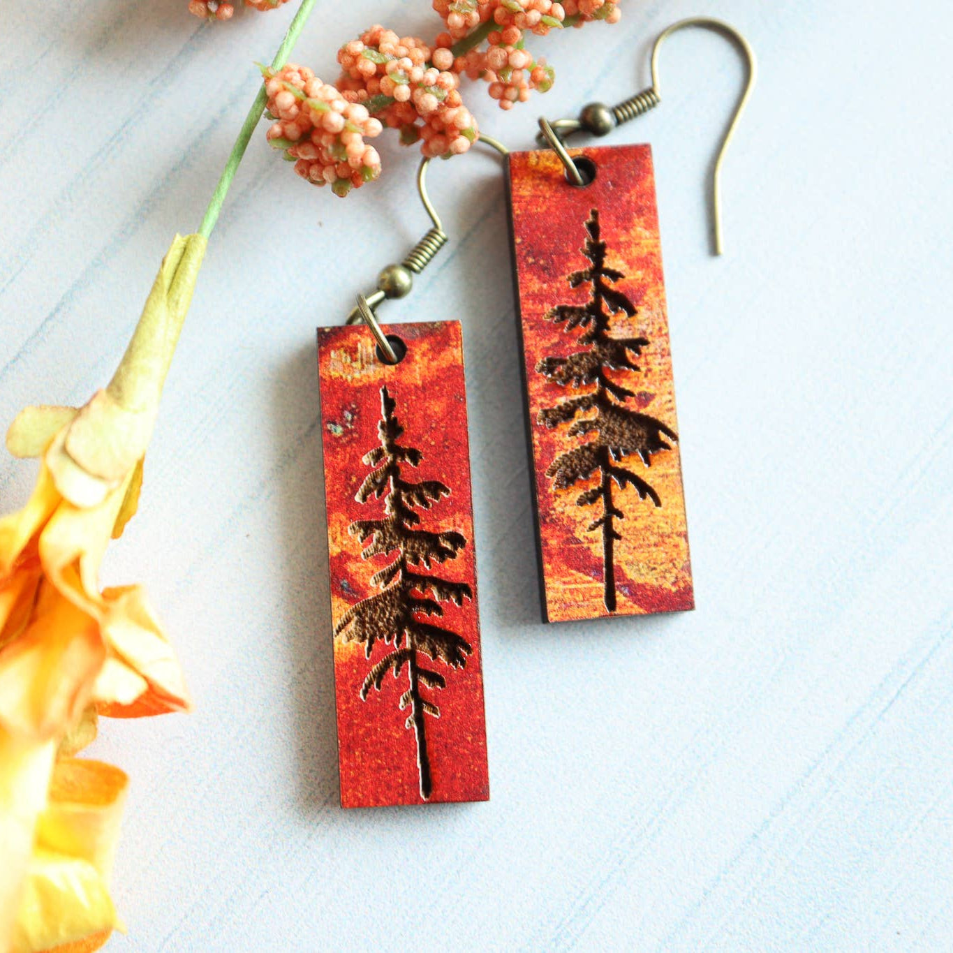 Pair of earrings with tree design on red background, surrounded by flowers.