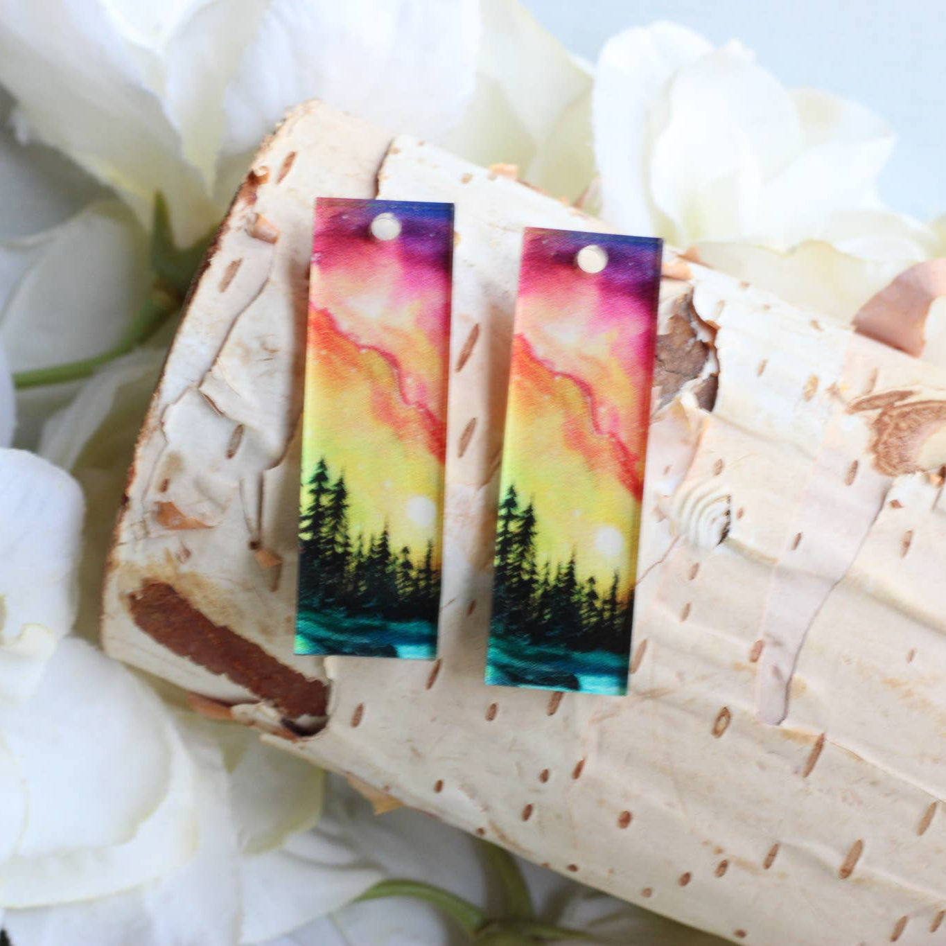 Colorful nature-themed earrings on a wooden surface with white flowers in the background