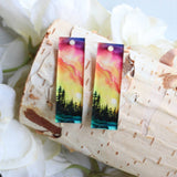 Colorful nature-themed earrings on a wooden surface with white flowers in the background
