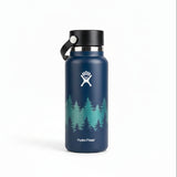 A Hydro Flask insulated water bottle in dark blue with a white forest tree graphic and the Hydro Flask logo, sitting on a wooden table with its box in the background.