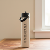 Beige water bottle with black cap and 'Montana' branding on a wooden surface.