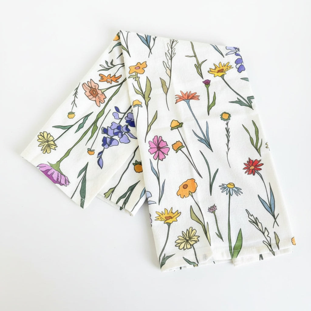 A folded white fabric napkin or tea towel with a colorful wildflower print, displayed on a white-washed wooden surface.