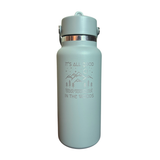A light sage green insulated water bottle with a matching lid and a silver metallic band. The front features a silver graphic of mountains, trees, and a sun, with the text 'IT'S ALL GOOD IN THE WOODS'.