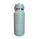 A light sage green insulated water bottle with a matching lid and a silver metallic band. The front features a silver graphic of mountains, trees, and a sun, with the text 'IT'S ALL GOOD IN THE WOODS'.