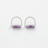A pair of silver hoop earrings with a small rectangular purple amethyst stone in the center, displayed on a white surface
