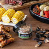 Assorted grilled foods including corn, chicken, and vegetables on a wooden surface with a jar of Applewood seasoning.