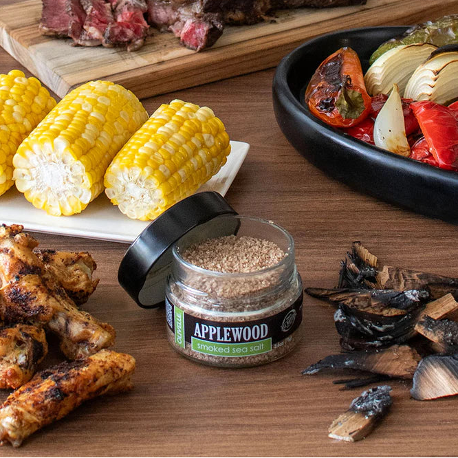 Assorted grilled foods including corn, chicken, and vegetables on a wooden surface with a jar of Applewood seasoning.
