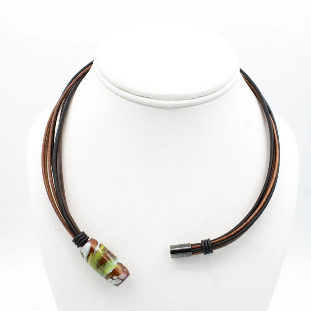 A multi-strand necklace with dark brown and black cords, featuring a large cylindrical glass lampwork bead with green, brown, and white patterns, displayed on a white mannequin bust.