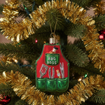 A Christmas tree ornament shaped like a barbecue apron hangs on a decorated tree branch. The apron is red on the upper portion with a green pocket area reading “BBQ BOSS,” and the lower green pockets contain barbecue tools such as tongs, a spatula, forks, and a bottle.