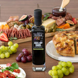 Bottle of Olivelle barrel aged dark balsamic vinegar on a wooden table with food items like bread, fruits, and salad.