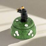 Decorative box with a black bear figurine on top, surrounded by white flowers on a green background.