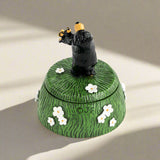 Decorative box with a black bear figurine on top, surrounded by white flowers on a green background.