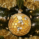 A gold Christmas ornament shaped like a honeycomb ball with three decorative bees and a hanging loop, displayed on a decorated Christmas tree with gold tinsel and warm lights.