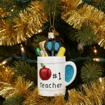 A white mug-shaped Christmas ornament hanging on a Christmas tree, decorated with a red apple, the text '#1 Teacher', and a blue glitter handle. The mug is filled with school supply decorations, including blue glitter scissors, a gold glitter pencil, and a yellow highlighter, and is surrounded by gold tinsel and green pine branches.