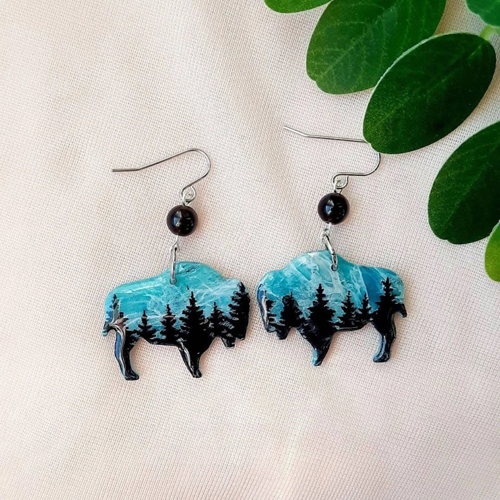 Bison-shaped earrings with turquoise and black design on a beige fabric background with green leaves.