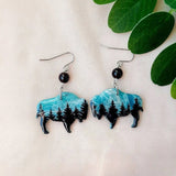 Bison-shaped earrings with turquoise and black design on a beige fabric background with green leaves.