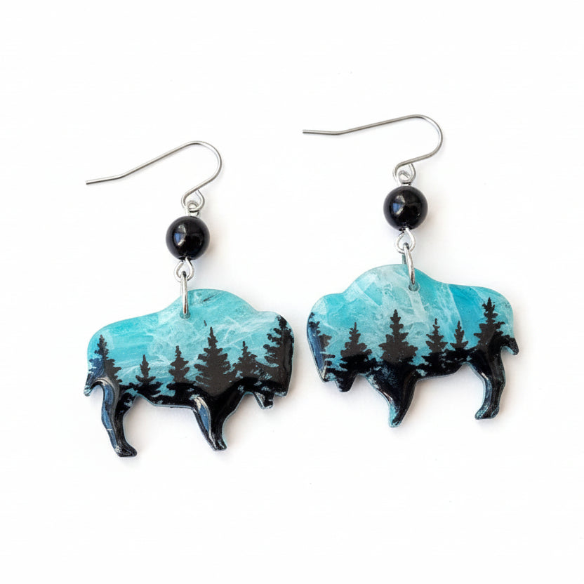 Bison-shaped earrings with turquoise and black design on a beige fabric background with green leaves.