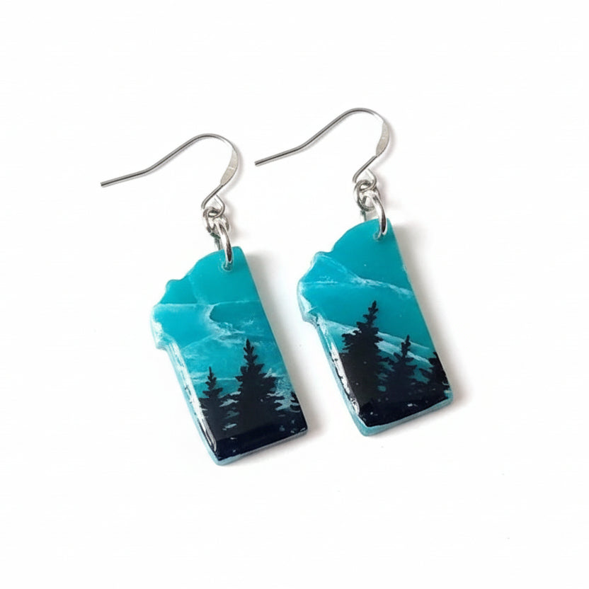 Teal and black landscape earrings on a white background with green leaves.