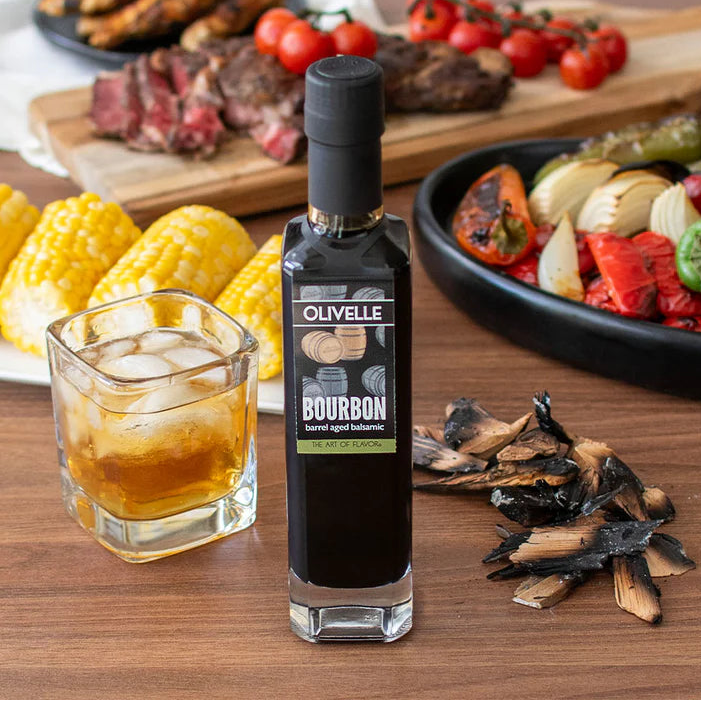 Bottle of Olivelle bourbon-aged balsamic vinegar on a wooden table with food and drinks.