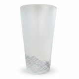 A frosted glass tumbler with a detailed gray mountain landscape illustration near the base, standing on a wooden surface with a dark teal tiled background.