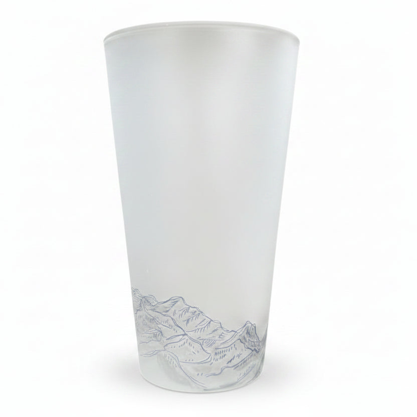 A frosted glass tumbler with a detailed gray mountain landscape illustration near the base, standing on a wooden surface with a dark teal tiled background.