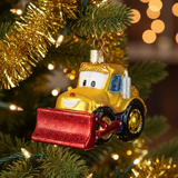 A gold Christmas tree ornament shaped like a cartoon snowplow truck with a red glitter front blade, hanging on a green Christmas tree with gold tinsel and warm bokeh lights in the background.