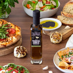 Bottle of Olivelle Caramelized Garlic oil on a table with pizza, salad, and bread.