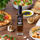 Bottle of Olivelle Caramelized Garlic oil on a table with pizza, salad, and bread.