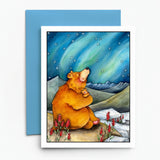 A square greeting card with a white border featuring a watercolor illustration of a brown bear hugging a small bear under a blue aurora sky, displayed on a white background with a blue envelope behind it.