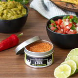 Olivelle Chipotle Lime rub & seasoning container with guacamole, salsa, tortilla chips, and limes on a wooden table.