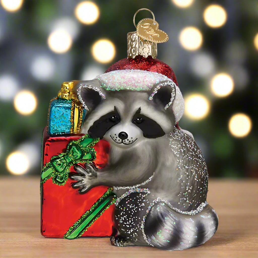Glass ornament of a raccoon wearing a Santa hat with presents on a white background