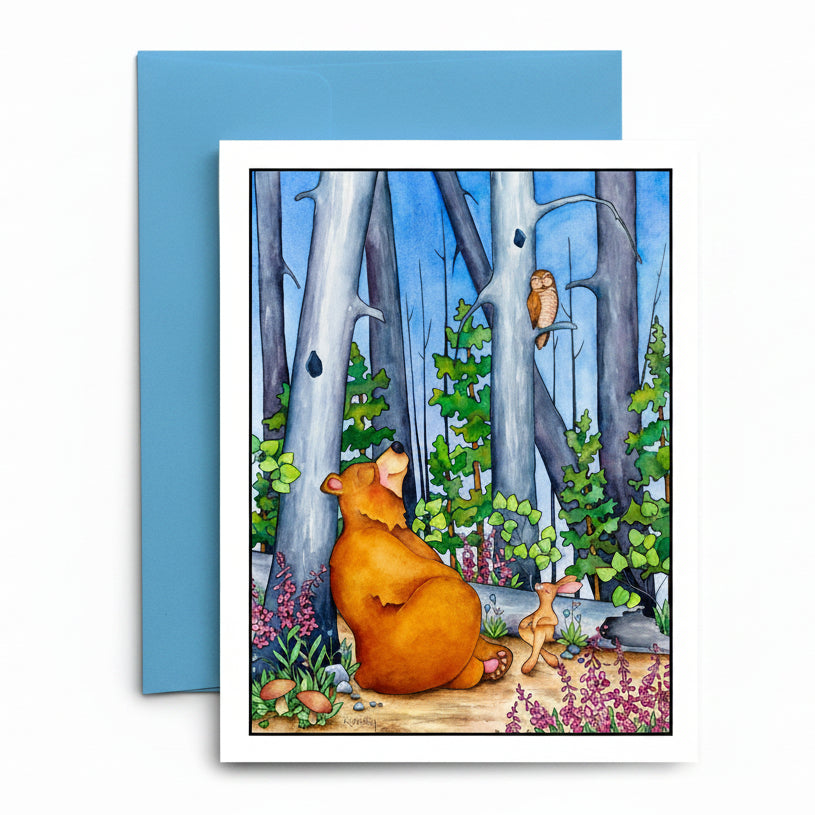 A square art print or greeting card with a white border showing a colorful forest illustration of a brown bear, a small deer, and an owl, placed in front of a blue envelope on a light wood surface.