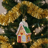 A gingerbread house Christmas ornament hanging from a green Christmas tree branch, surrounded by gold tinsel and warm string lights.