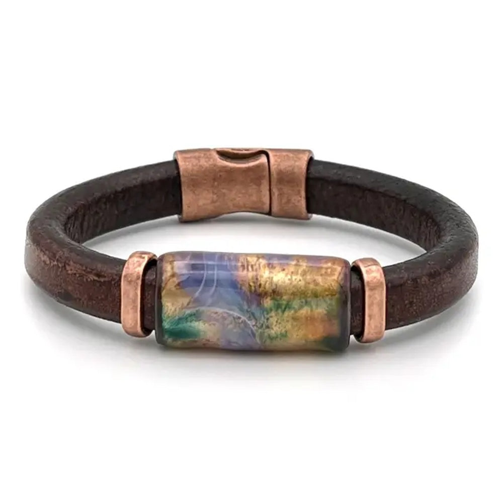 A dark brown leather bracelet with a central rectangular glass bead featuring a colorful landscape design in gold, blue, and green, accented by copper-toned metal caps and a magnetic clasp.