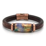A dark brown leather bracelet with a central rectangular glass bead featuring a colorful landscape design in gold, blue, and green, accented by copper-toned metal caps and a magnetic clasp.