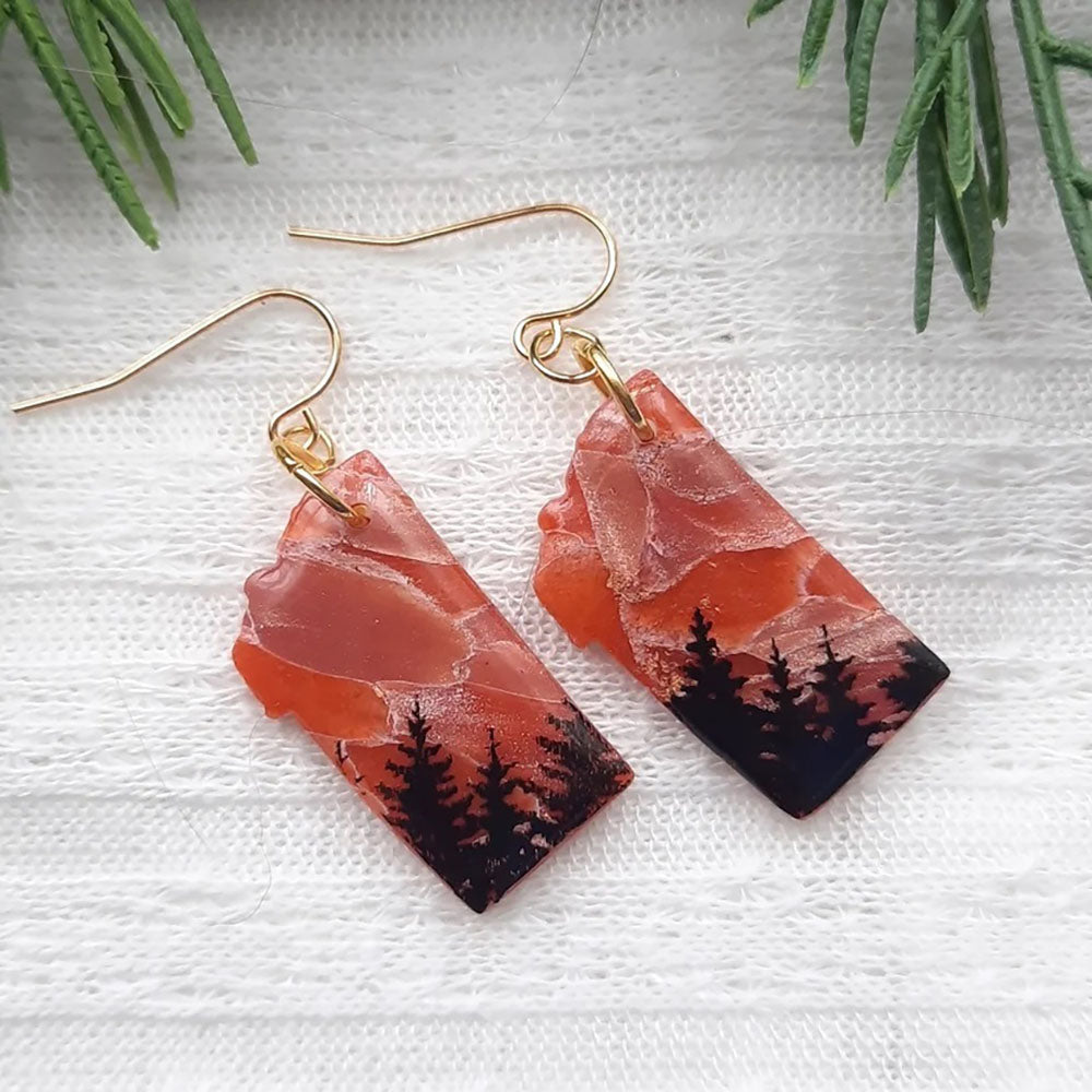 Pair of earrings with nature-themed design on a textured white surface with greenery.