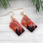 Pair of earrings with nature-themed design on a textured white surface with greenery.