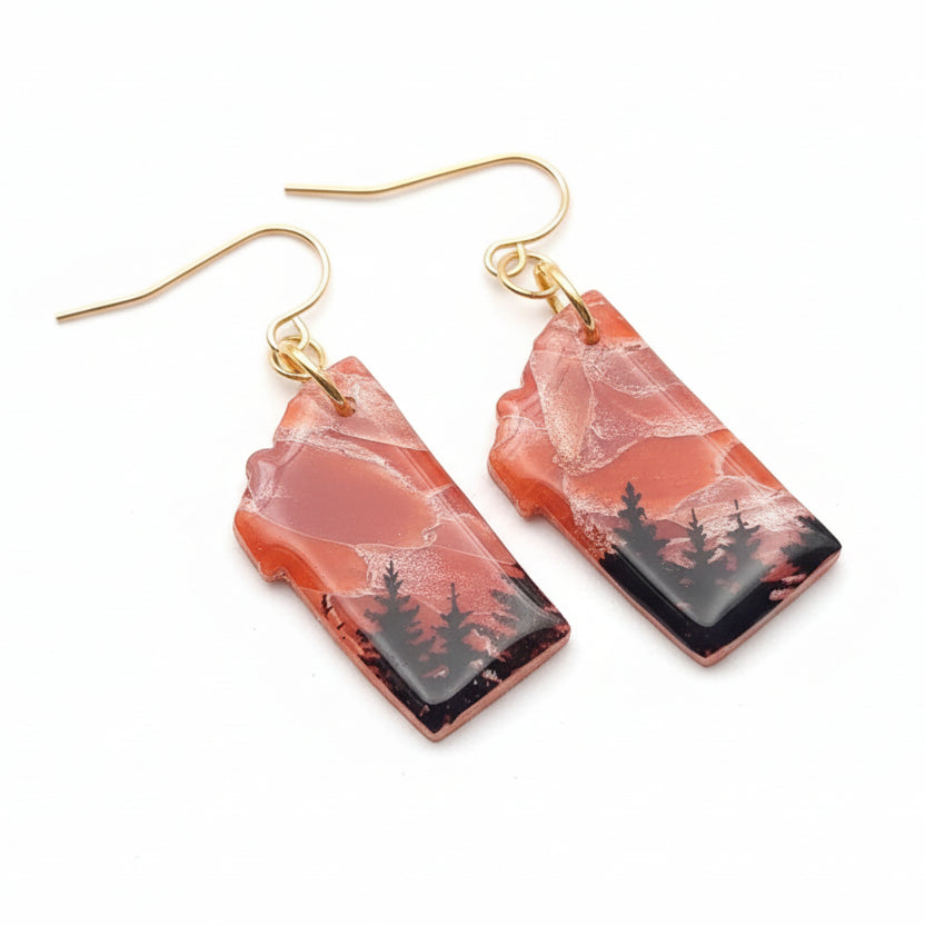 Pair of earrings with nature-themed design on a textured white surface with greenery.