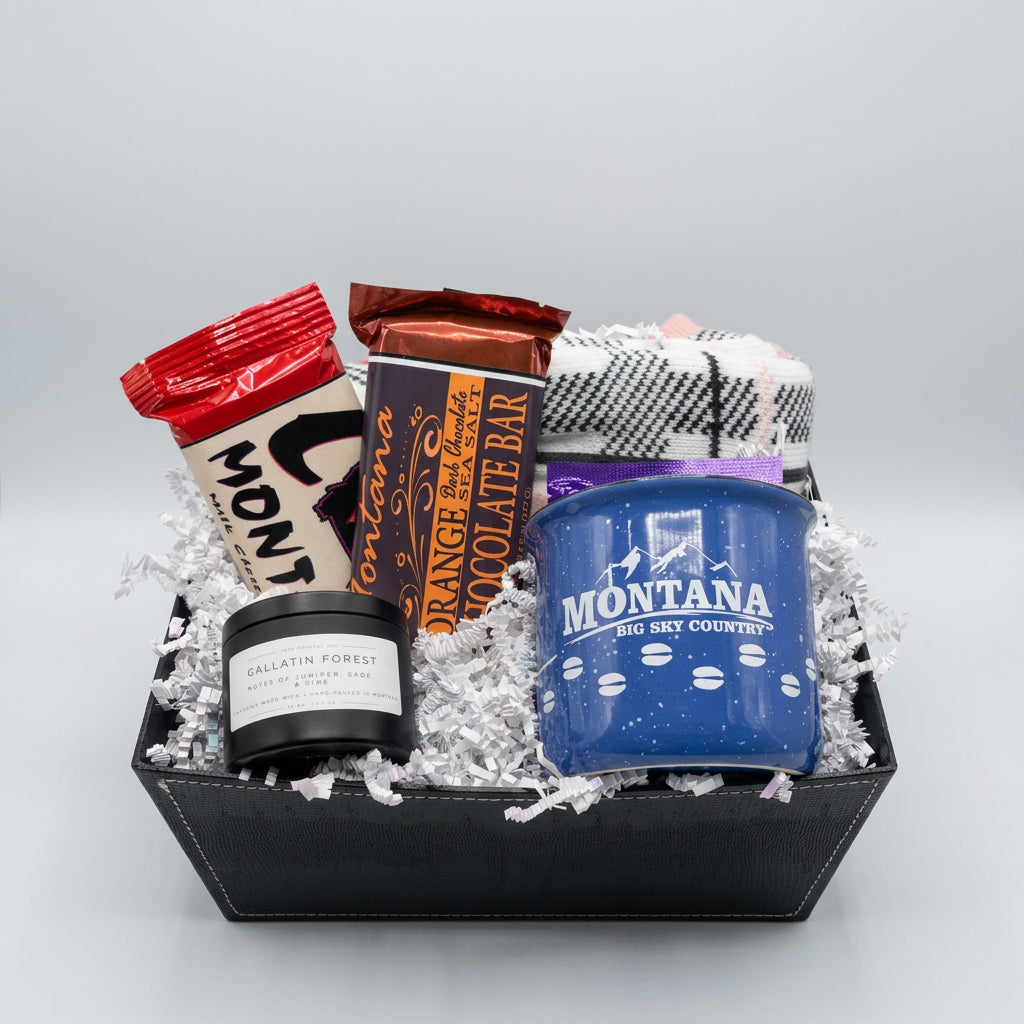 A curated gift basket in a black tray, filled with shredded white paper and containing multiple items: a blue enamel mug with Montana branding, a black candle jar labeled Gallatin Forest, two wrapped chocolate bars, a plaid fabric item, and a purple wrapped set of tea bags