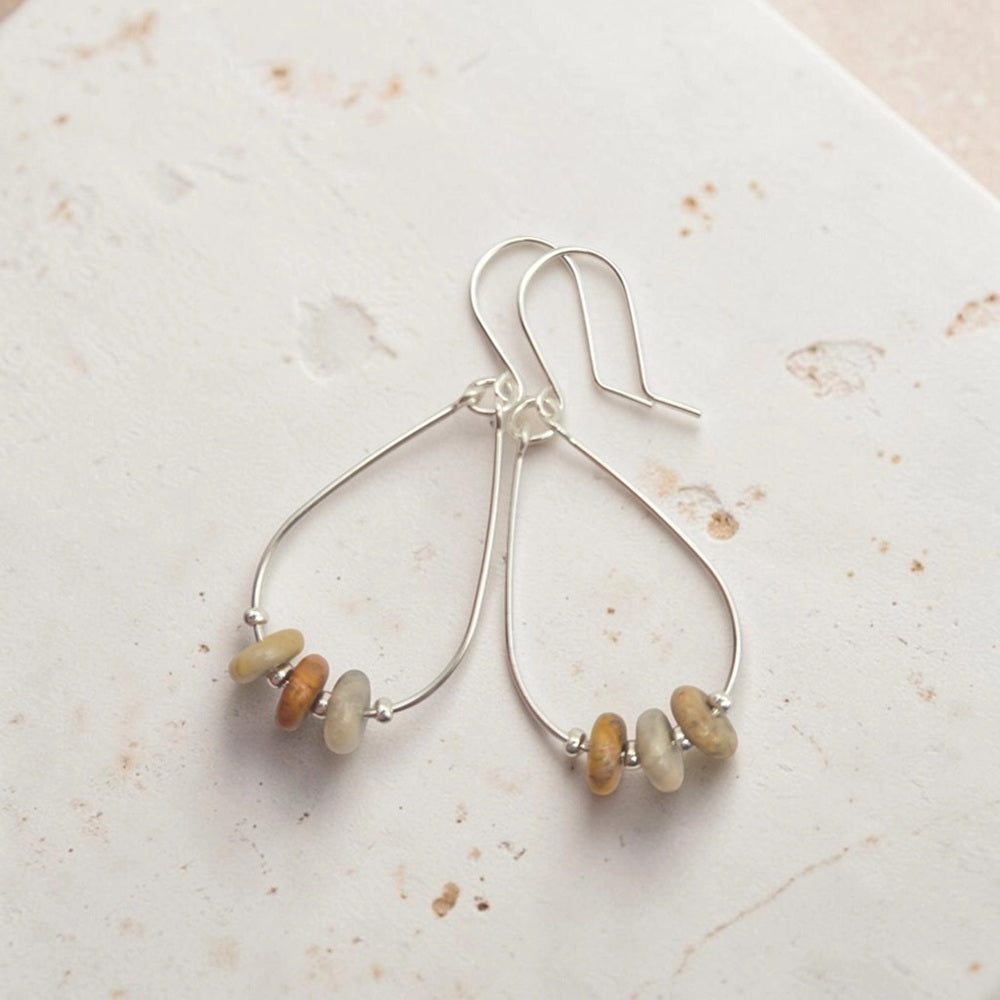A pair of silver teardrop dangle earrings with small, irregular natural stone beads in muted beige, brown, and cream, displayed on a textured off-white surface.