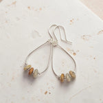 A pair of silver teardrop dangle earrings with small, irregular natural stone beads in muted beige, brown, and cream, displayed on a textured off-white surface.