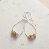 A pair of silver teardrop dangle earrings with small, irregular natural stone beads in muted beige, brown, and cream, displayed on a textured off-white surface.