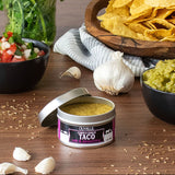 Olivelle corn & garlic taco rub on a wooden table with tortilla chips, guacamole, and salad.