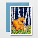 A square art print or card featuring a watercolor-style scene of a large brown bear and a small rabbit in a flower field at night, placed in front of a blue envelope on a light wood surface.