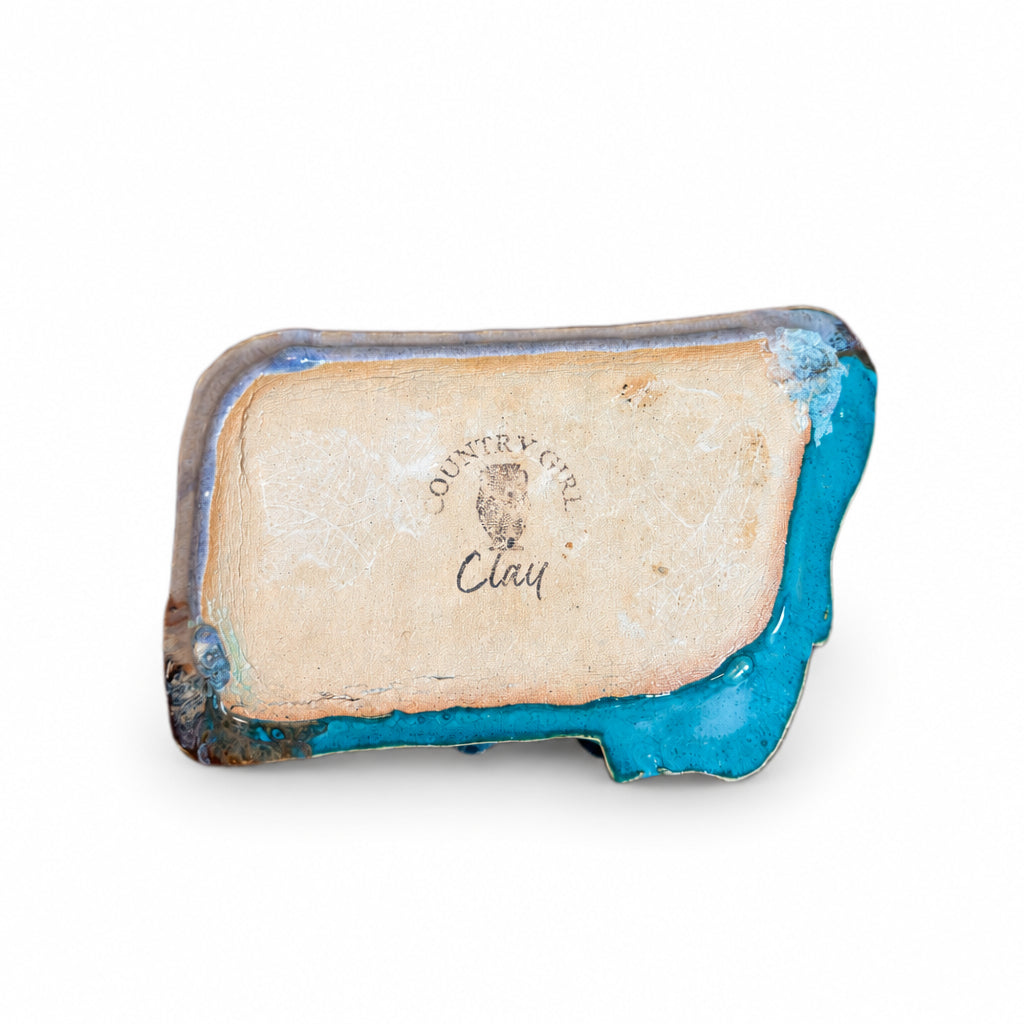A handmade ceramic clay dish with a rustic turquoise blue glazed edge and a beige sandy center, resting on a dark wood surface against a light green wall. The center features a stamped logo reading 'Country Clay Clay'.